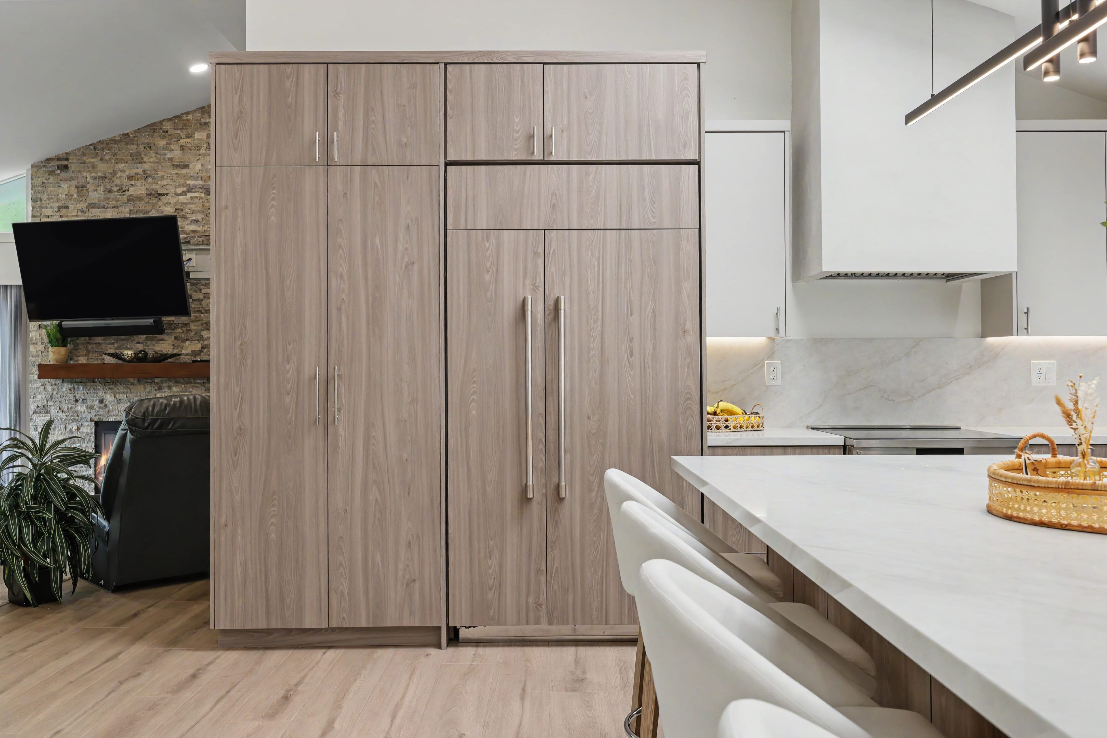 Tall wood-grain kitchen pantry cabinets and a built-in refrigerator next to a white marble kitchen island with white bar stools.