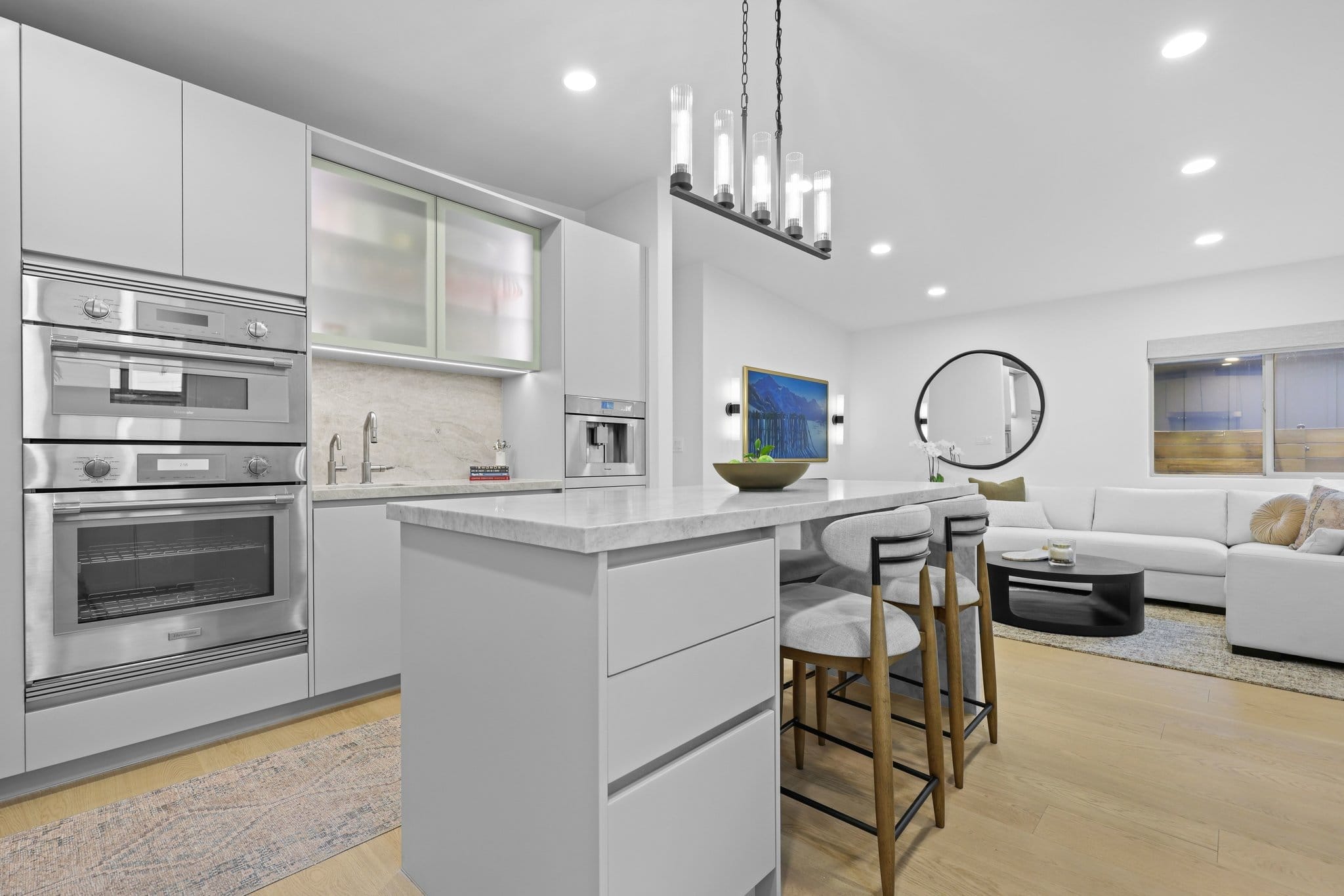 Modern white kitchen with quartz waterfall island, stainless wall ovens and open living room