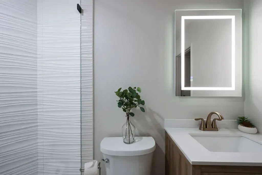 Compact powder room with textured white tile, wood vanity and backlit mirror
