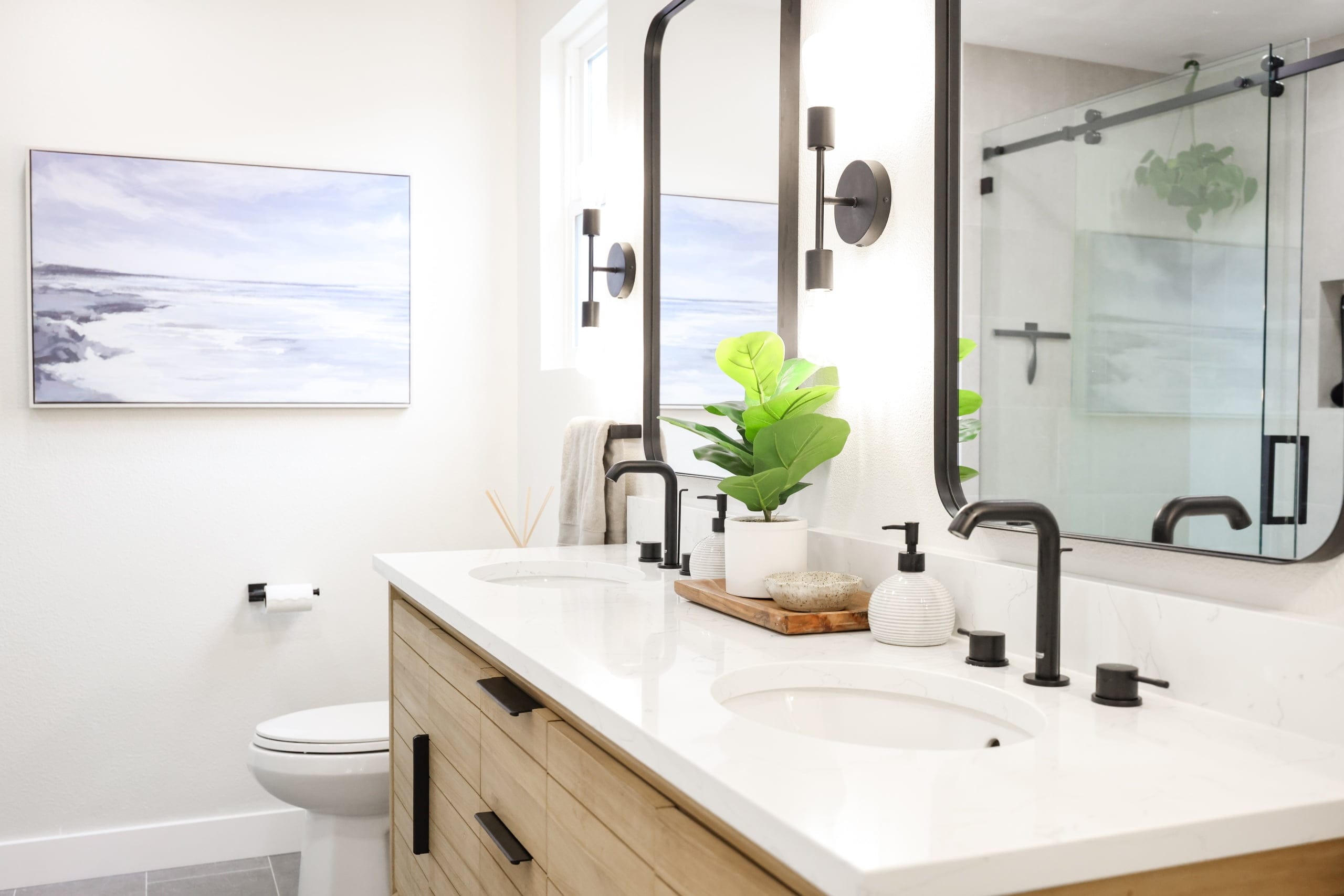 light wood double vanity with coastal art, black framed mirrors and glass shower