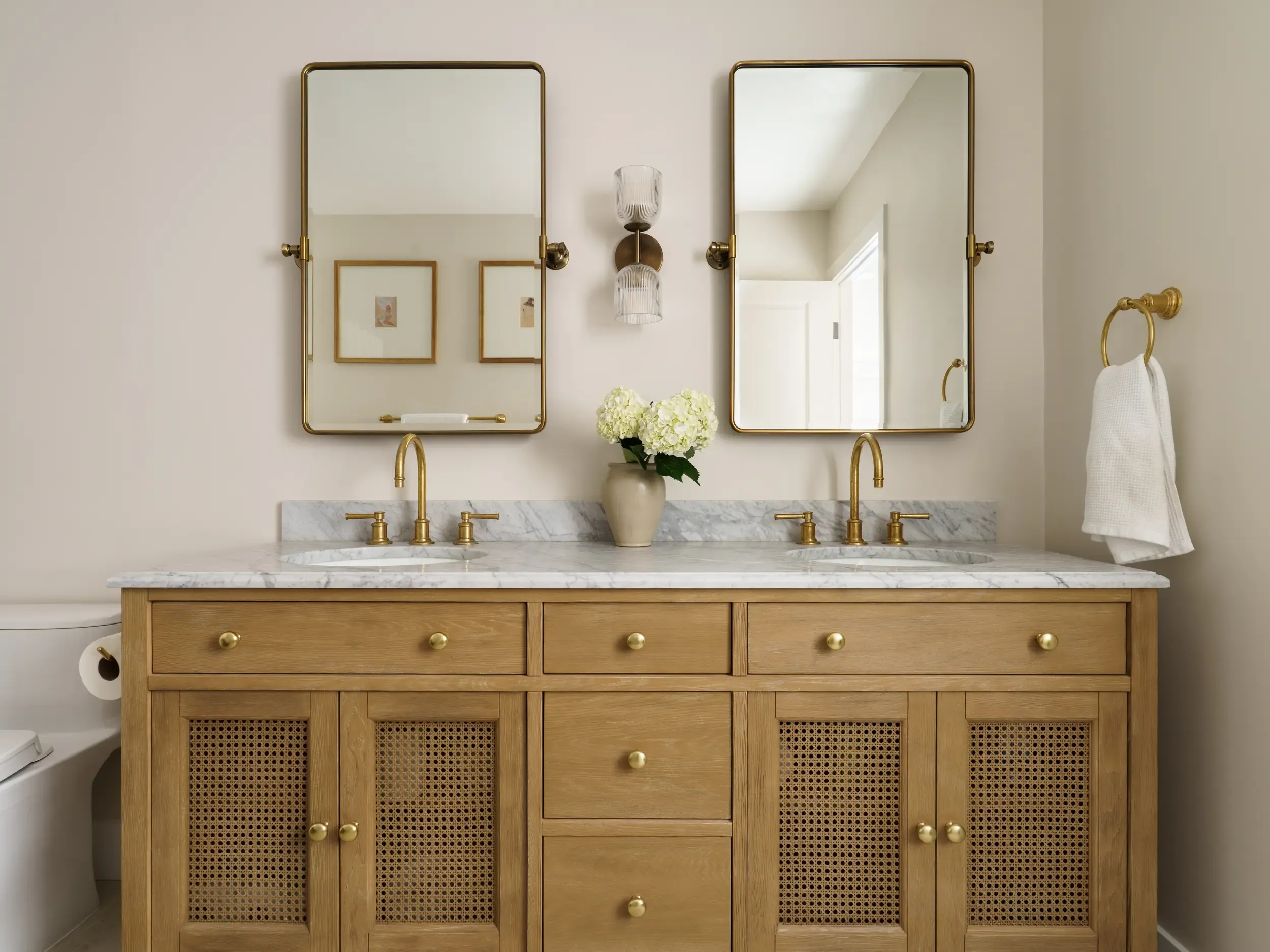 Remodeled bathroom with oak cane-front double vanity, marble countertop, and brass pivot mirrors