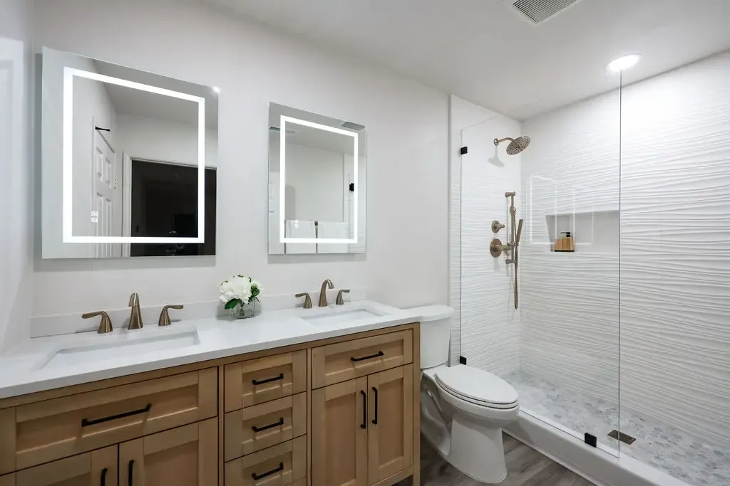 Bathroom with natural wood double vanity, lit mirrors and walk-in tiled shower