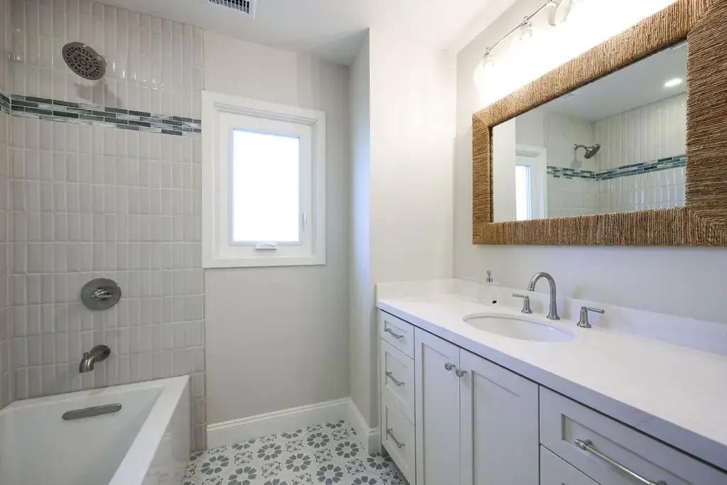 Narrow bathroom with tiled tub surround, white vanity and woven framed mirror